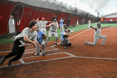 An infield drill in the dome: a batter at the plate, a catcher in full gear crouched behind, and fielders reacting across the dirt