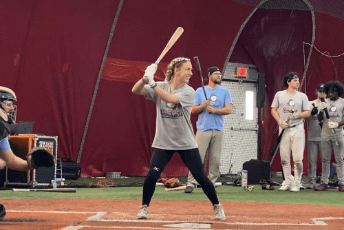 A player in a Snowballs shirt loads up in her batting stance in the indoor cage while teammates wait their turn behind her