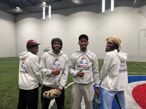 Four players in Snowballs hoodies stand shoulder to shoulder on the indoor turf, gloves in hand, smiling at the camera