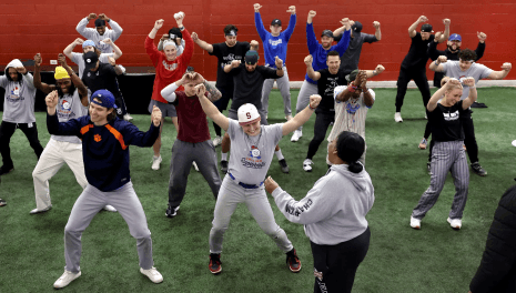 A large group of players in a wide stance with fists raised, running through choreography led by a coach on indoor turf