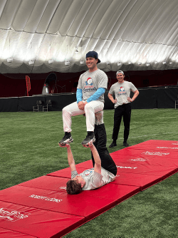 Two performers practising a partner balance on a red mat: one lies on their back supporting the other, who stands upright on their hands